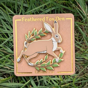 May include: A rectangular enamel pin featuring a brown hare leaping through a green leafy wreath. The pin has a gold border and the text "Feathered Fox Den" and "Enamel Pin". The pin is set against a backdrop of green grass.