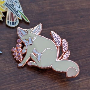 May include: A rose gold enamel pin featuring a white fennec fox sitting amongst purple lavender flowers. The fox has green eyes and a pink nose.