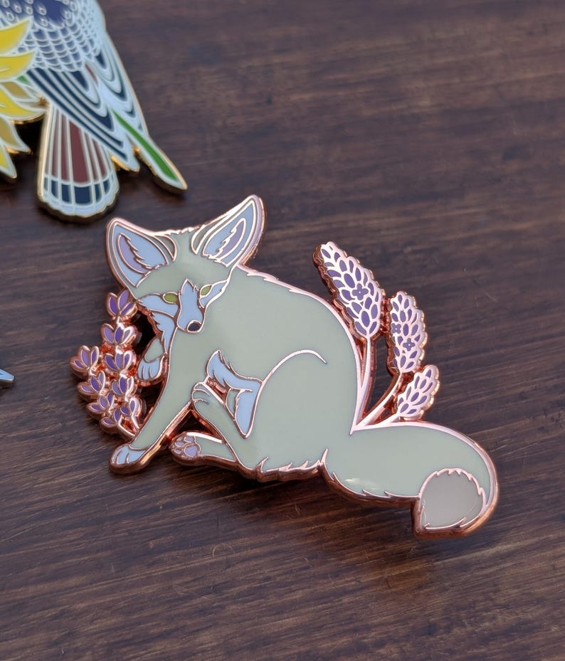 May include: A rose gold enamel pin featuring a white fennec fox sitting amongst purple lavender flowers. The fox has green eyes and a pink nose.