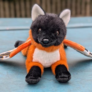 May include: A plush toy of a fox-like creature with black fur on its head and paws, orange fur on its body, and white accents. It has large, stylized wings with orange, black, and white patterns. The toy is lying on a blue surface.