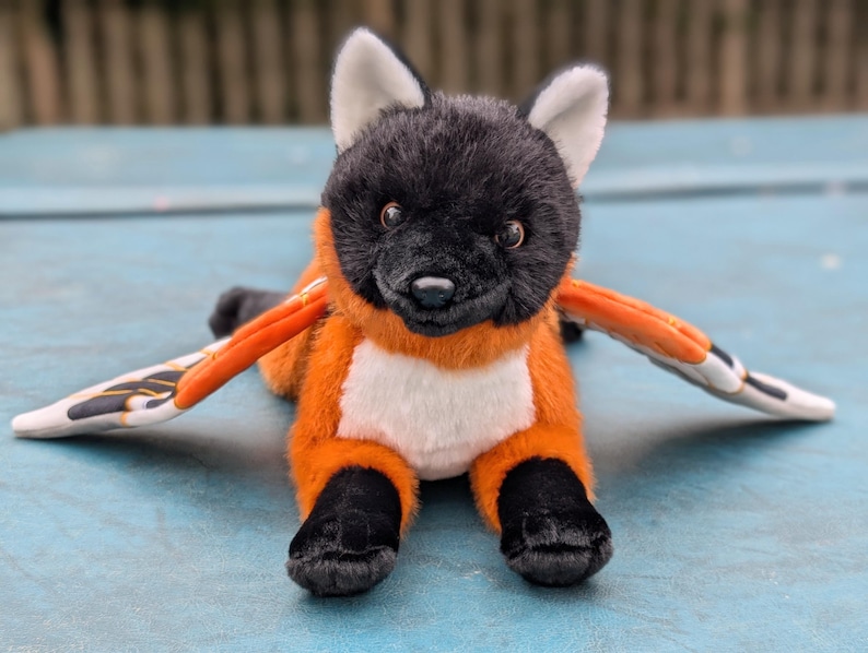 May include: A plush toy of a fox-like creature with black fur on its head and paws, orange fur on its body, and white accents. It has large, stylized wings with orange, black, and white patterns. The toy is lying on a blue surface.