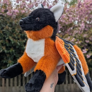 May include: A plush toy creature with a black head and paws, orange chest and sides, and white belly. It has large wings with orange, black, and white details. The toy is held up against a blurred background of pink flowers.