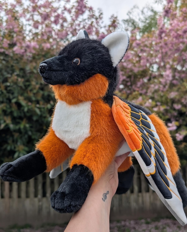 May include: A plush toy creature with a black head and paws, orange chest and sides, and white belly. It has large wings with orange, black, and white details. The toy is held up against a blurred background of pink flowers.