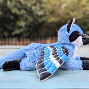 May include: A plush blue jay toy with black and white accents. The bird has a detailed wing design with blue, black, and white feathers. The toy is lying down on a light blue surface, showcasing its soft, plush texture.
