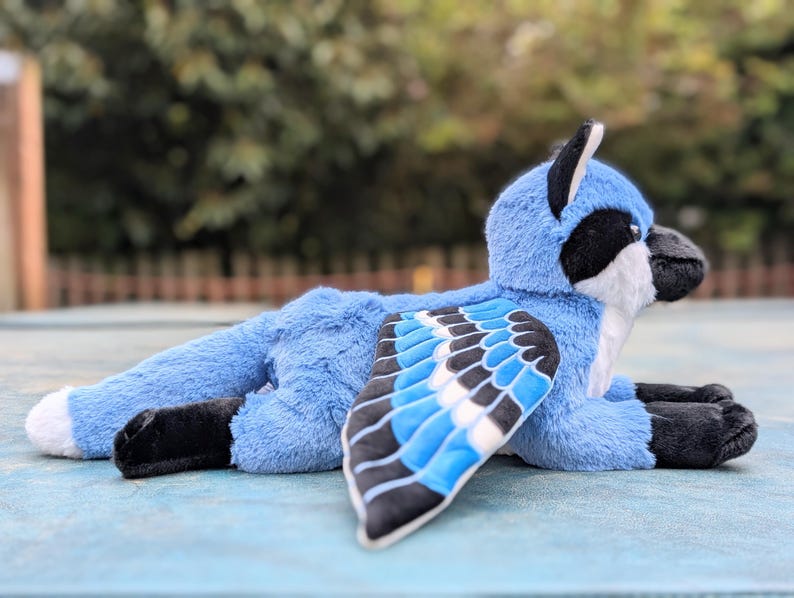 May include: A plush blue jay toy with black and white accents. The bird has a detailed wing design with blue, black, and white feathers. The toy is lying down on a light blue surface, showcasing its soft, plush texture.