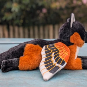 May include: A plush toy fox with black, orange, and white fur, and a wing-like design on its side. The fox is lying down, with its paws and tail visible. The wings feature a pattern of black, orange, and white.
