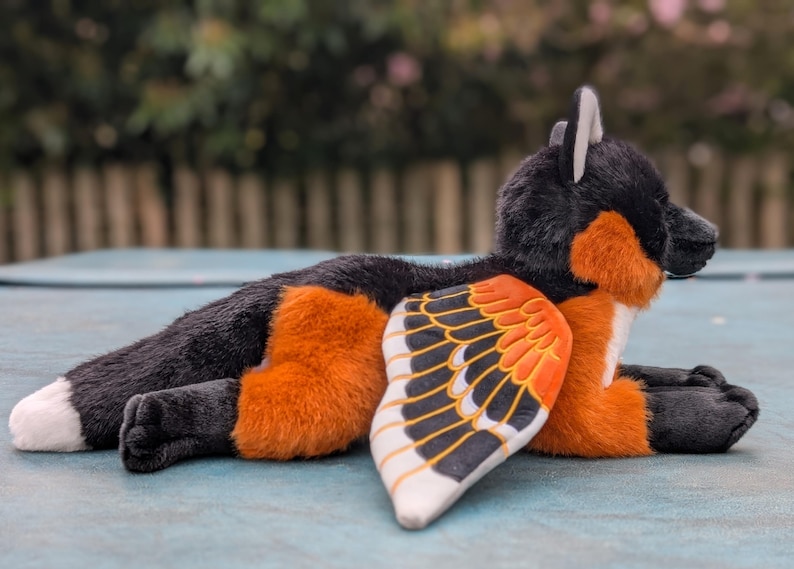 May include: A plush toy fox with black, orange, and white fur, and a wing-like design on its side. The fox is lying down, with its paws and tail visible. The wings feature a pattern of black, orange, and white.
