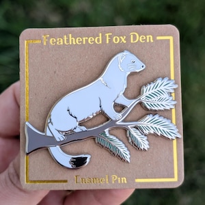 May include: A silver enamel pin featuring a white weasel sitting on a branch with green leaves. The pin is on a brown card with the text "Feathered Fox Den" and "Enamel Pin".