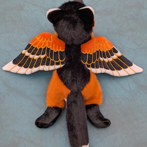 May include: A plush toy fox with black fur, orange pants, and wings. The wings are orange with black and white details. The fox has a long, black tail with a white tip. The toy is lying on a teal surface.
