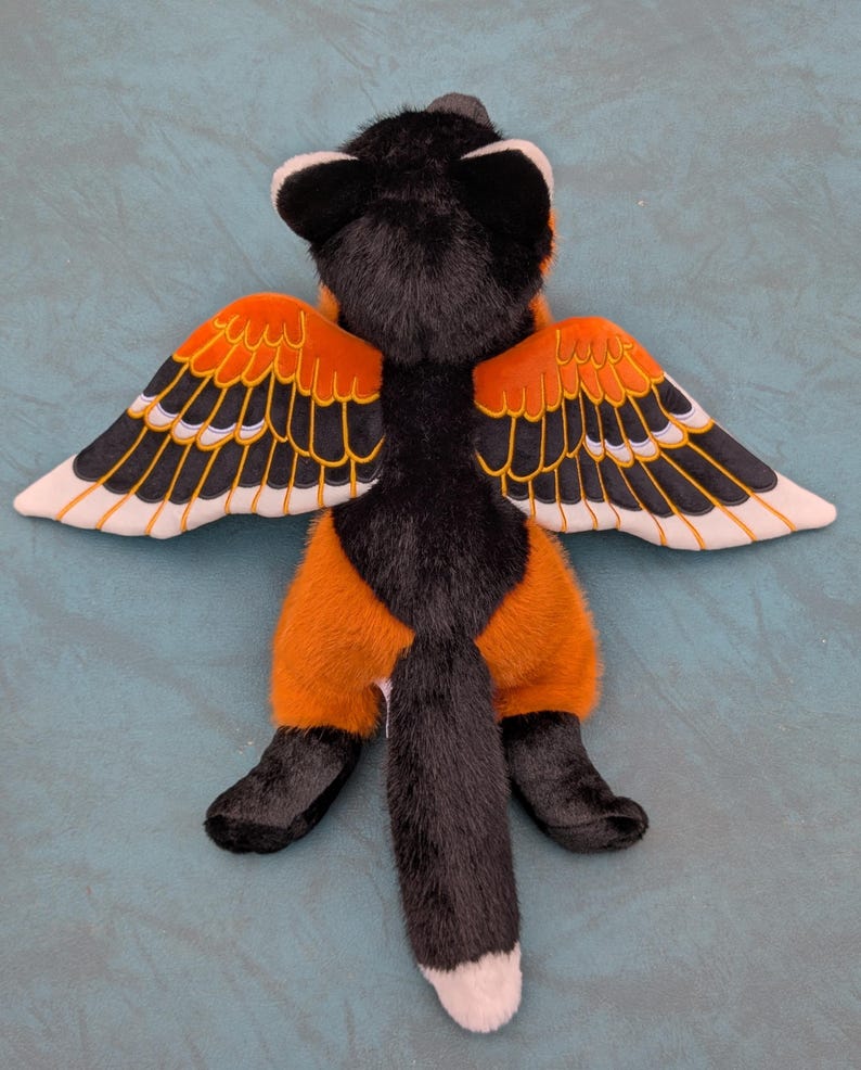 May include: A plush toy fox with black fur, orange pants, and wings. The wings are orange with black and white details. The fox has a long, black tail with a white tip. The toy is lying on a teal surface.