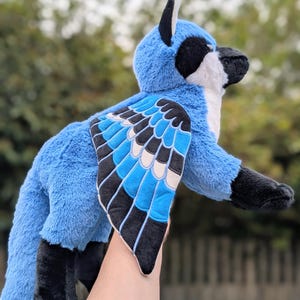 May include: A plush blue jay bird with black and white accents. The stuffed animal has a fluffy texture and detailed wings with blue, black, and white patterns. The bird's beak is black, and it has a white chest.