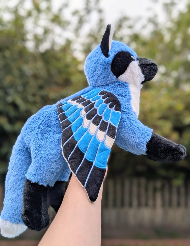 May include: A plush blue jay bird with black and white accents. The stuffed animal has a fluffy texture and detailed wings with blue, black, and white patterns. The bird's beak is black, and it has a white chest.