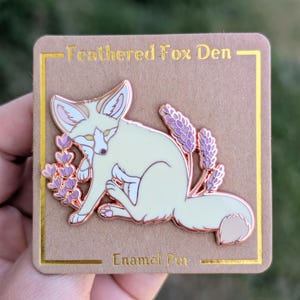 May include: A light yellow enamel pin of a fennec fox sitting with its ears back, surrounded by purple lavender flowers. The pin is on a brown card with the text "Feathered Fox Den" and "Enamel Pin".
