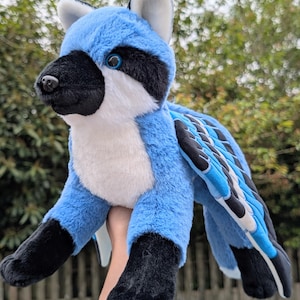 May include: A plush toy creature with blue, black, and white fur. The toy has a raccoon-like mask, blue wings, and black paws. The creature is held up against a blurred green background.