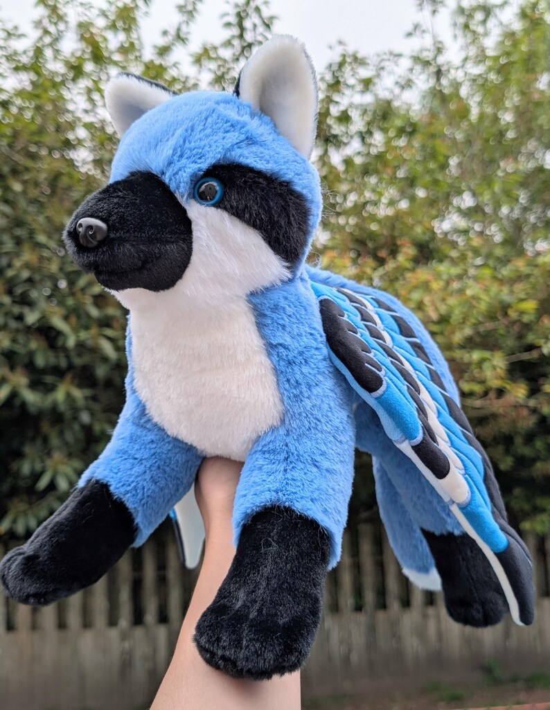 May include: A plush toy creature with blue, black, and white fur. The toy has a raccoon-like mask, blue wings, and black paws. The creature is held up against a blurred green background.