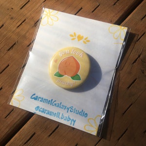 May include: A yellow button with a peach illustration and the text "you look peachy!"  The button is packaged in a clear plastic bag with a white card that says "CaramelGalaxyStudio @caramell.baby"