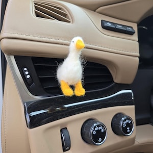 May include: A white felted goose with a yellow beak and feet is positioned in a car's air vent. The car's interior features tan and black elements, including control knobs and vents. The goose is the central object.