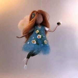 May include: A handmade fairy doll with red hair, a blue dress decorated with white and yellow flowers, and black leggings. The fairy holds a small bee. The doll has white wings and is suspended.