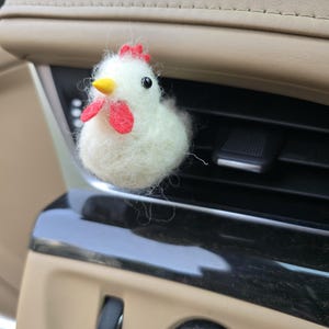 May include: A white felt chicken with a red comb and wattle, and a yellow beak. The chicken is an air vent accessory, designed to clip onto a car's air vent. The chicken is a decorative item.