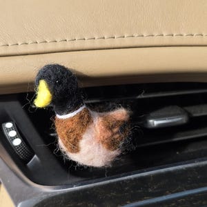May include: A felt duck ornament with a black head, brown and white body, and yellow beak. The duck is positioned in a car's air vent. This decorative item adds a playful touch to the vehicle's interior.