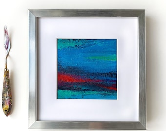 Puede incluir: Una pintura abstracta enmarcada con un esquema de color azul, verde y rojo. La pintura está en un marco plateado y está colgada en una pared blanca.