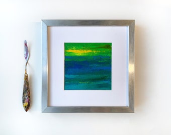 Puede incluir: Una pintura abstracta enmarcada con pinceladas horizontales en tonos de verde, azul y amarillo. La pintura está en un marco plateado.