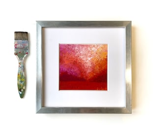 Puede incluir: Una pintura abstracta enmarcada con un esquema de color rojo, naranja y rosa. La pintura está en un marco plateado y sobre un fondo blanco.