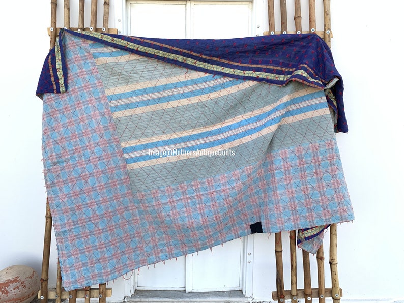 May include: A blue and white kantha quilt with a striped pattern and a checkered pattern. The quilt is hanging on a bamboo rack.
