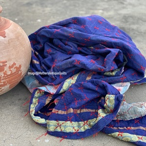 May include: A blue kantha quilt with red embroidery and gold trim. The quilt is folded and lying on a concrete surface next to a terracotta pot.
