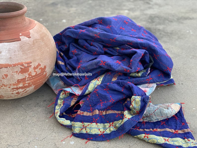 May include: A blue kantha quilt with red embroidery and gold trim. The quilt is folded and lying on a concrete surface next to a terracotta pot.