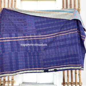 May include: A blue kantha quilt with a red and white geometric pattern. The quilt is folded and hanging on a wooden rack. The quilt has a light blue and white border.