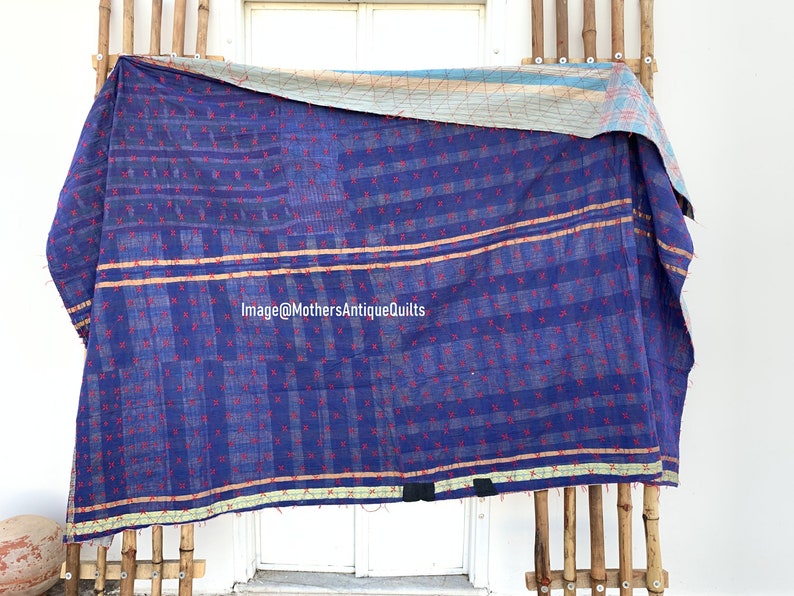 May include: A blue kantha quilt with a red and white geometric pattern. The quilt is folded and hanging on a wooden rack. The quilt has a light blue and white border.