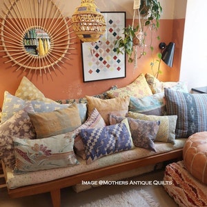 Op de afbeelding: Een houten bank met een verscheidenheid aan kleurrijke, gepatroneerde kussens. De bank staat in een kamer met een lichtbruine muur en een wit tapijt. De tekst "IMAGE @MOTHERS ANTIQUE QUILTS" is zichtbaar op de bank.