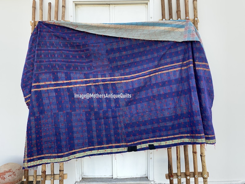 May include: A blue kantha quilt with red stitching and a light brown border. The quilt is hanging on a wooden rack in front of a white door. Image@MothersAntiqueQuilts