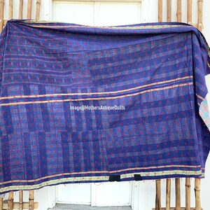 May include: A blue kantha quilt with a red and white geometric pattern. The quilt has a gold and green border and is hanging on a bamboo rack.