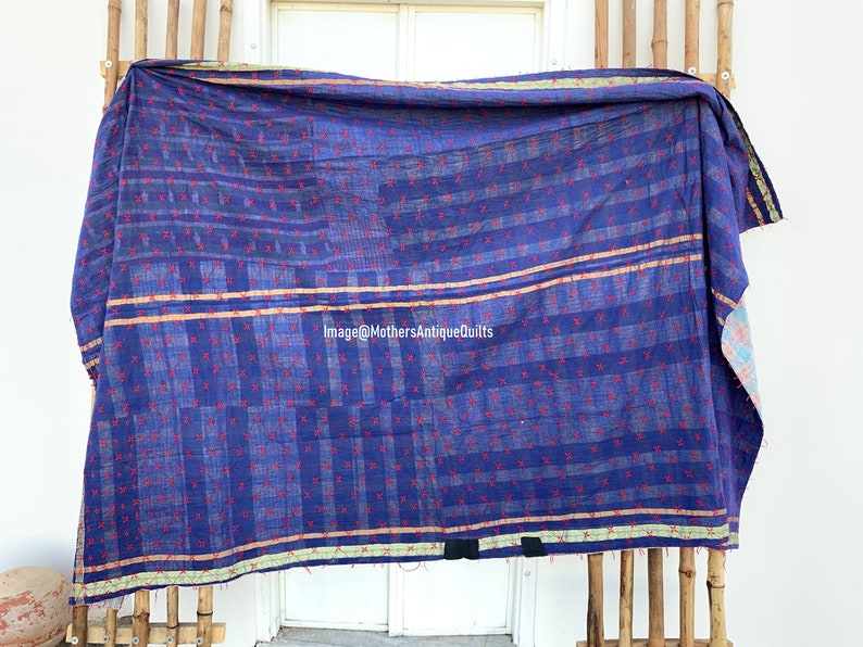 May include: A blue kantha quilt with a red and white geometric pattern. The quilt has a gold and green border and is hanging on a bamboo rack.