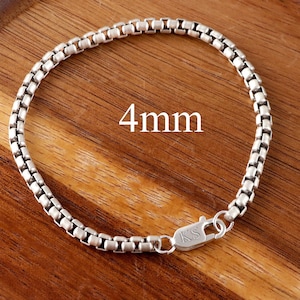 May include: A silver-toned box chain bracelet with a lobster claw clasp featuring the engraved letters KS. The chain links have a square profile and the text 4mm is visible in the centre of the circular arrangement of the bracelet.