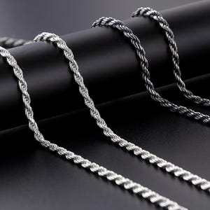 May include: Three silver-coloured rope chain necklaces are displayed against a black background. The chains have a twisted design, with one appearing darker than the others. The image highlights the texture and detail of the jewellery.