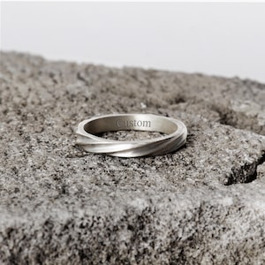 Könnte beinhalten: Ein silberfarbener Metallring mit einem gedrehten, spiralförmigen Banddesign. Die Innenfläche des Rings zeigt das Wort Custom, das in einer klaren serifenlosen Schrift eingraviert ist. Das Band hat ein mattes Finish und eine fließende Form.