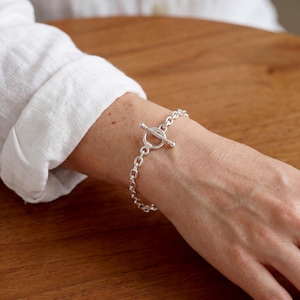 Puede incluir: Una pulsera de cadena plateada con cierre de barra y argolla. La pulsera está formada por una serie de eslabones ovalados interconectados, con un anillo circular y una barra perpendicular que asegura la cadena alrededor de la muñeca.