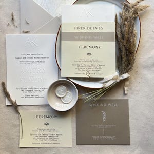 Puede incluir: Elegante juego de invitaciones de boda con texto en relieve y diseño minimalista. Incluye tarjetas de invitación, tarjetas RSVP y un sobre. La paleta de colores es neutra con tonos crema, beige y blanco. Decorado con flores secas.