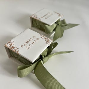 Puede incluir: Dos cajas de regalo blancas con lazos de cinta verde. Las cajas están decoradas con un patrón floral y el texto "Pamela & Chad" en letras doradas.