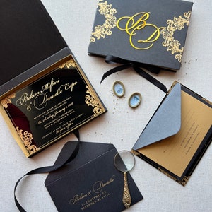 Puede incluir: Elegantes invitaciones de boda negras con detalles dorados. El conjunto incluye una caja, un sobre y una tarjeta. Las invitaciones presentan los nombres "Bekim & Danielle" y los detalles del evento.