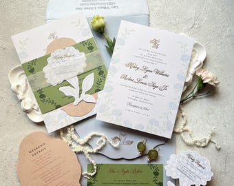May include: Wedding invitation suite featuring floral designs and elegant typography. Includes invitation cards, RSVP cards, and envelopes in shades of green, peach, and blue. A decorative floral embellishment and a sprig of flowers add to the aesthetic.