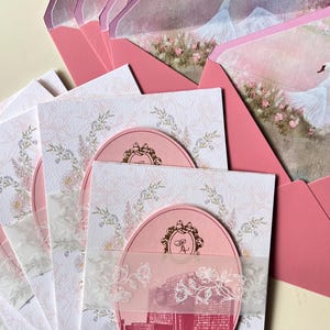 May include: Pink wedding invitations with a floral design and a pink oval with a city scene. The invitations have a sheer overlay with a floral design and are paired with pink envelopes featuring a swan illustration.