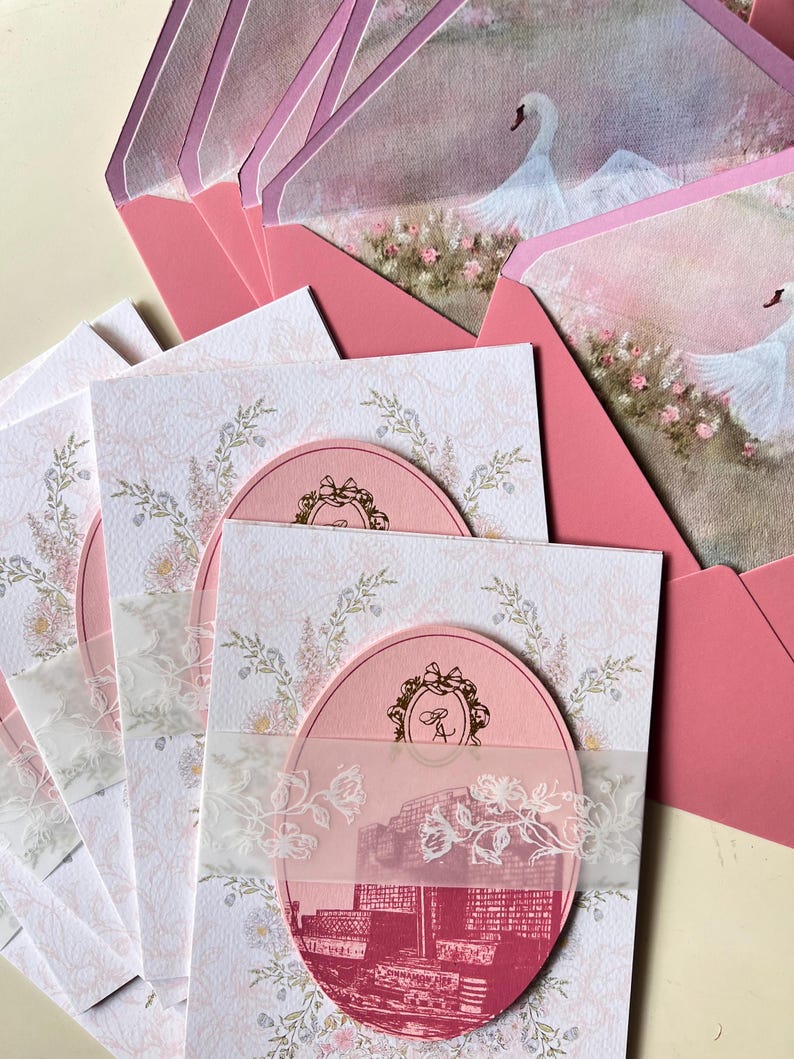 May include: Pink wedding invitations with a floral design and a pink oval with a city scene. The invitations have a sheer overlay with a floral design and are paired with pink envelopes featuring a swan illustration.