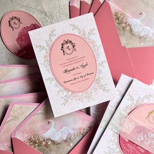 May include: Pink wedding invitations and envelopes. The invitation features an oval design with floral accents and the text "Ramasha & Angelo". The envelopes have a swan illustration. A matching round card is also visible.