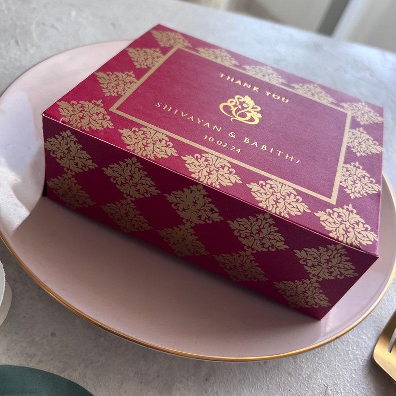 Puede incluir: Una caja de agradecimiento roja y dorada con un patr&oacute;n decorativo. La caja dice "Gracias" y "Shivayan & Babith/ 10.02.24".