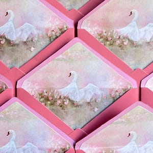 May include: Pink envelopes with a swan illustration. Each envelope features a painting of a white swan with outstretched wings, set against a soft pink and floral background. The envelopes are arranged in a repeating pattern.