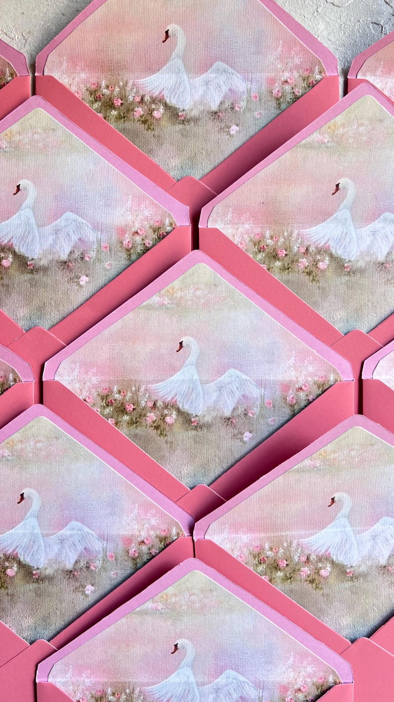 May include: Pink envelopes with a swan illustration. Each envelope features a painting of a white swan with outstretched wings, set against a soft pink and floral background. The envelopes are arranged in a repeating pattern.